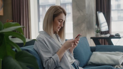 woman sitting on couch in modern home using phone and smiling texting message watching contents or...