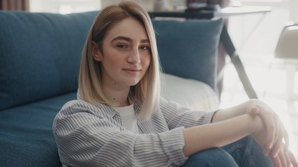 Portrait of cheerful young caucasian woman sitting on the floor near couch looking to the camera smiling enjoying having fun at modern room no stress concept