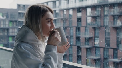 Fototapeta premium Close up view of cheerful young caucasian woman in white hoodie stays in balcony drink hot coffee or tea relax enjoy breathing fresh air healthy freezy snow falls in march morning no stress concept