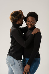 smiling african american women in black turtlenecks hugging isolated on grey