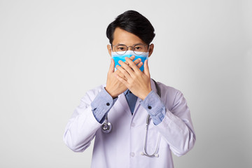 attractive asia man doctor wearing surgical mask and hand close mouth gesture standing in gray wall background. Concept Preventive measures against corona virus.