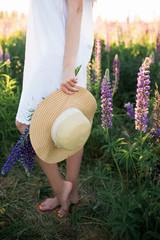 The girl in the field holds a straw hat and a bouquet of lupins in her hands.
