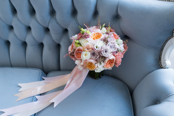 Delicate wedding bridal bouquet with white, orange and gray-purple flowers with long pink ribbons. The bouquet lies on a blue sofa