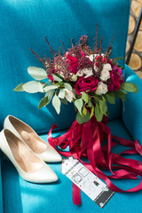 White and red wedding bridal bouquet, along with wedding shoes and other accessories of the bride lie on a blue armchair. Wedding details