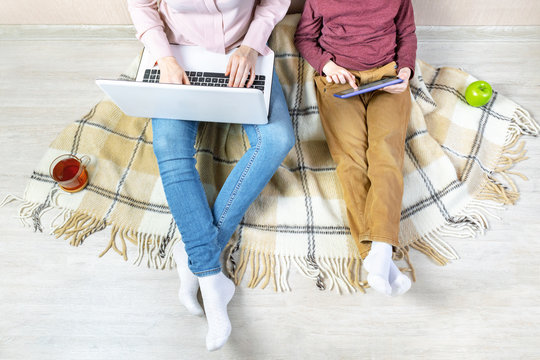 Mom works on a laptop and her son plays on a tablet, remote work from home, freelance, quarantine. A young woman and a preschooler boy are sitting together on a plaid on the floor in the apartment.
