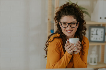 Portrait of attractive young woman with curly hair and eyeglasses who enjoying drinking coffee during the break from work. Attractive young woman is posing in front of camera.