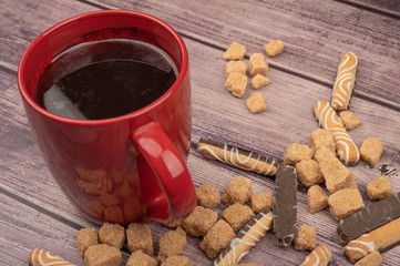 A red ceramic tea mug, cookie sticks with chocolate and white icing, and pieces of brown cane sugar on a wooden background. Close up.