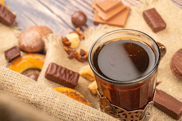 A faceted glass of tea in a vintage Cup holder, pieces of chocolate, bagels and chocolate gingerbread on a background of rough homespun fabric. Close up.