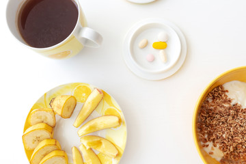 Small bowl with medicines, yellow bowl with white yogurt and flakes, yellow saucer with apple slices and banana, yellow cup with black hot tea