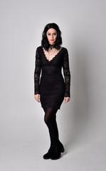 Naklejka premium Portrait of a goth girl with dark hair wearing black lace dress and boots. Full length standing pose on a studio background.