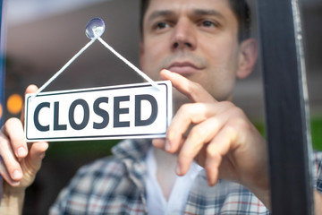 Small Business Owner With Serious Expression Putting Up Closed Sign During Recession Or Health...
