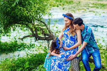 Naklejka premium Pregnant woman mother to be in wreath and blue dress sitting with her daughter and husband near scenic river in sunset time in summer. Pregnancy and healthy organic close to nature lifestyle.