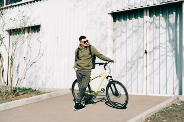 a young man in khaki sportswear holds a Bicycle standing next to him, against the background of a light hangar on a Sunny spring day