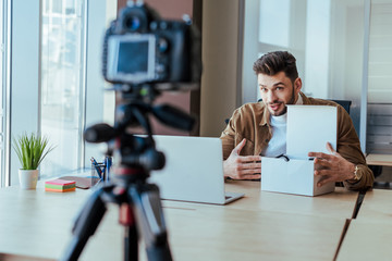 Selective focus of blogger pointing at box near laptop at table in front of digital camera