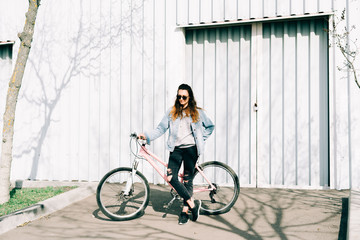 a young stylish girl holds a pink Bicycle standing next to her, against the background of a light hangar on a Sunny spring day