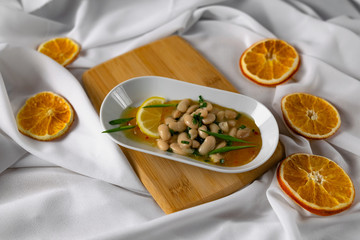 Marinated white beans in their own juice on an oval plate with Thai pineapple sauce and green onions. Decorated with a slice of lemon and dried oranges on a wooden Board with a snow white fabric