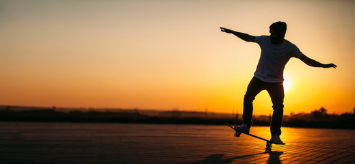Skater hipster man riding on a board on a city street during sunset wearing jeans and white t shirt with a hat. Empty space for text