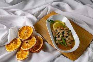 Marinated white beans in their own juice on an oval plate with Thai pineapple sauce and green onions. Decorated with a slice of lemon and dried oranges on a wooden Board with a snow white fabric