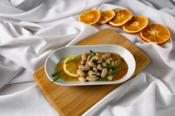 Marinated white beans in their own juice on an oval plate with Thai pineapple sauce and green onions. Decorated with a slice of lemon and dried oranges on a wooden Board with a snow white fabric