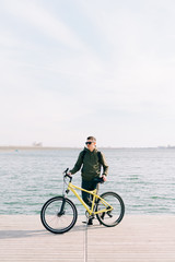 a young man in khaki sportswear and glasses stands on a pontoon near the lake and holds a yellow Bicycle near him on a Sunny spring day