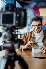 Selective focus of blogger with opened box smiling and looking at digital camera at table in coworking space