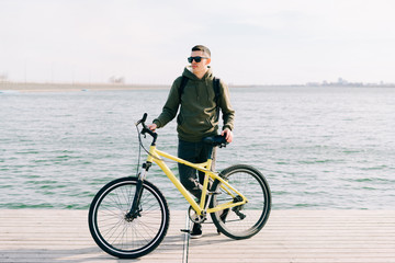 Fototapeta premium a young man in khaki sportswear and glasses stands on a pontoon near the lake and holds a yellow Bicycle near him on a Sunny spring day