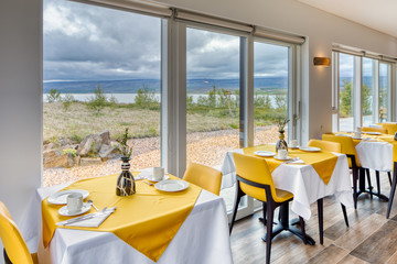 Elegant and minimalist restaurant in a Scandinavian hotel with fantastic views of the countryside and mountains. Tables with yellow and white tablecloths with breakfast cutlery. Large windows.