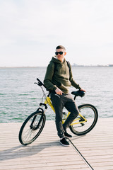 Fototapeta premium a young man in khaki sportswear and glasses stands on a pontoon near the lake and holds a yellow Bicycle near him on a Sunny spring day