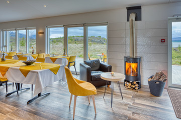 Elegant and minimalist restaurant in a Scandinavian hotel with fantastic views of the countryside and mountains. Yellow and white tablecloths. Large windows, wood-burning stove and leather sofa