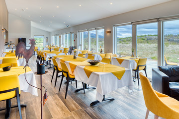 Elegant and minimalist restaurant in a Scandinavian hotel with fantastic views of the countryside and mountains. Tables with yellow and white tablecloths with breakfast cutlery. Large windows.
