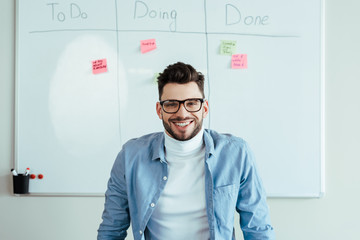 Scrum master looking at camera and smiling near white board with stickers and spreadsheet with to do, doing, done lettering © LIGHTFIELD STUDIOS
