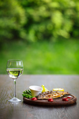 Roasted fish with vegetables on the wooden board with a glass of white wine on the wooden table, green background, vertical, side view