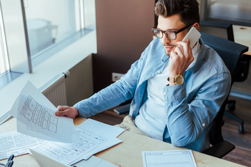 High angle view of IT worker holding papers, talking on smartphone and smiling at table in coworking space