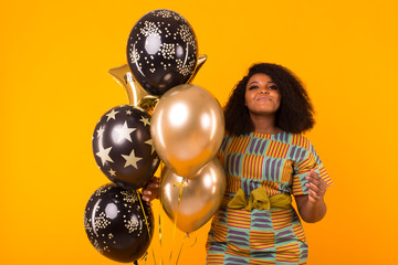 Naklejka premium Holidays, birthday party and fun concept - Portrait of smiling young African-American young woman looking sweet on yellow background holding balloons.