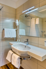 Interior of bathroom in minimalist modern style with mirror, sink on stone stand, towells, hairdresser, shampoo, gel and wooden tiles. Soft cream and white color