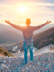 Man stands on the top of a mountain with his hands up. In the background, sunset and the sea, winding serpentine road below. Freedom travel concept