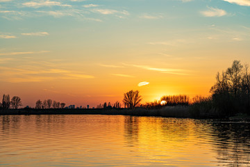 The setting sun is barely visible through the trees along the lake Zoetermeerse Plas, Zoetermeer, Netherlands
