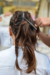Fototapeta premium Super styling. Rear view closeup of a hairdresser braiding her clients hair in trendy weave plait while open-air festival