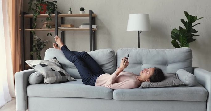 Relaxed young woman lying on sofa in living room using smart phone. Lazy happy millennial lady customer surfing social media, shopping in app, playing mobile game, texting sms looking at cell at home.
