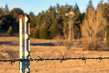 close up of barbed wire fence on a T post in foreground a rural autumn fall setting with windmill...