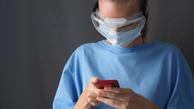 Female Nurse In Mask Use A Smartphone On A Gray Background, A Female Doctor Is Holding A Phone. Covid-19 Pandemic Coronavirus.