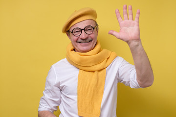 Senior man in glasses and yellow hat and scarf saying hello, waving a hand.