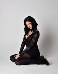 Portrait of a goth girl with dark hair wearing black lace dress and boots. Full length sitting...