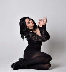 Portrait of a goth girl with dark hair wearing black lace dress and boots. Full length sitting...