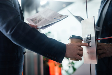 Cropped view of businessman with newspaper and paper cup pushing button of elevator