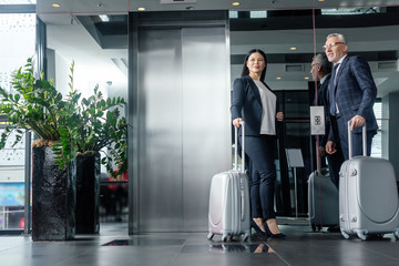 smiling businessman and asian businesswoman with travel bags standing near elevator