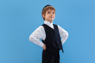 Posing confident, cute. Portrait of a young orthodox jewish boy isolated on blue studio background....