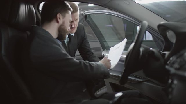 Auto dealership customer is signing a cintract sitting in a new car. Сar salesman is telling interested buyer about luxurious car. The customer is reading the contract in motor show.