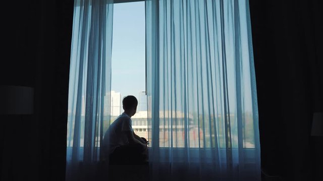 A young handsome young man sits at the window behind a curtain and looks at the landscape of a large metropolis.