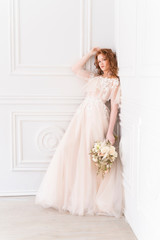 Beautiful natural redhead girl bride, with nude makeup, wearing a white dress, holds a wedding bouquet in her hands, standing an the wall in a light interior.